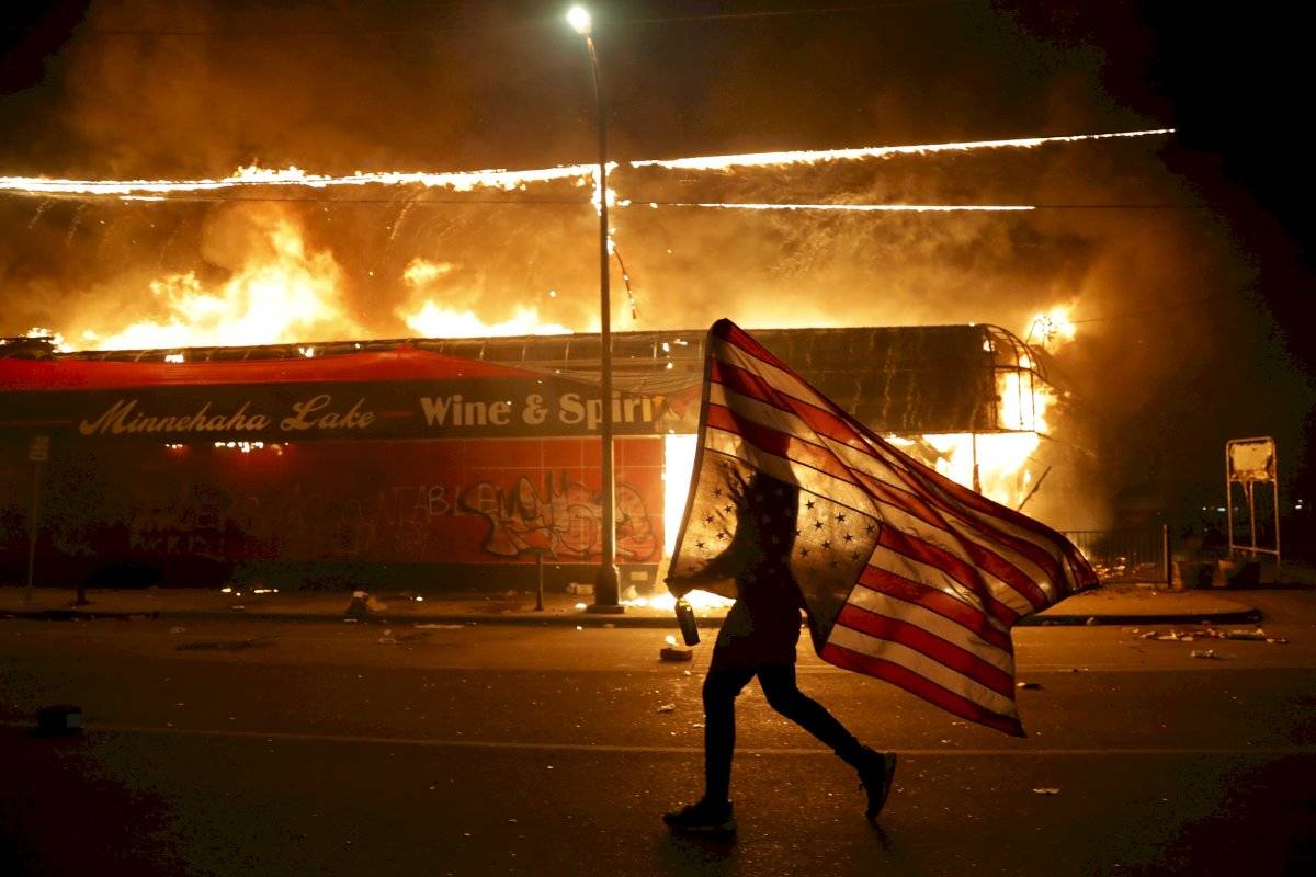 Un manifestante lleva una bandera estadounidense al revés, un signo de angustia, junto a un edificio en llamas el jueves 28 de mayo de 2020 en Minneapolis